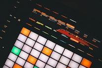 An electronic music production controller featuring illuminated pads and digital display with sound frequency controls. Various buttons and sliders with labels like 'Mute', 'Solo', and 'Quantize' are visible.