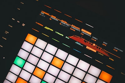 An electronic music production controller featuring illuminated pads and digital display with sound frequency controls. Various buttons and sliders with labels like 'Mute', 'Solo', and 'Quantize' are visible.