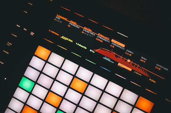 An electronic music production controller featuring illuminated pads and digital display with sound frequency controls. Various buttons and sliders with labels like 'Mute', 'Solo', and 'Quantize' are visible.