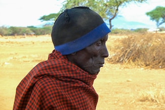 An elderly person wearing a checkered red and black cloth draped over their shoulders, with a black and blue knit hat on their head. The background features a dry, open landscape with sparse vegetation and a few trees, suggesting a rural or arid environment.