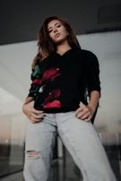 A person is standing confidently with hands in their pockets. They are wearing a black sweatshirt with a floral design and light-colored ripped jeans. The setting seems to be outdoors or in a modern architectural space with glass elements.