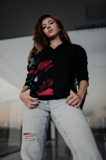 A person is standing confidently with hands in their pockets. They are wearing a black sweatshirt with a floral design and light-colored ripped jeans. The setting seems to be outdoors or in a modern architectural space with glass elements.