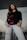 A person is standing confidently with hands in their pockets. They are wearing a black sweatshirt with a floral design and light-colored ripped jeans. The setting seems to be outdoors or in a modern architectural space with glass elements.