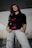 A person is standing confidently with hands in their pockets. They are wearing a black sweatshirt with a floral design and light-colored ripped jeans. The setting seems to be outdoors or in a modern architectural space with glass elements.