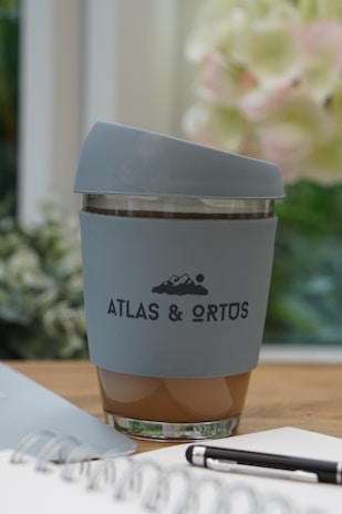 A reusable cup with a grey silicone sleeve and matching lid, displaying the logo 'ATLAS & ORTUS' along with a simple mountain design. The cup appears to contain a beverage, possibly coffee. It is placed on a wooden table indoors, with a blurry floral arrangement in the background. A notebook and pen are partially visible in the foreground.