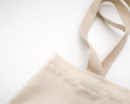A section of a beige canvas tote bag is visible, showcasing the stitching details and one handle extending upwards on a plain white background. The natural texture of the fabric is evident.