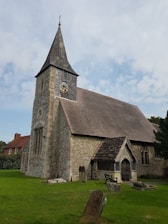 A historic stone church with a prominent steeple and a clock on the tower. The building features arched windows and a sloped roof. The foreground includes a well-maintained lawn with several old gravestones, adding a rustic and tranquil atmosphere. The surrounding area has some greenery, and part of a red-brick house is visible to the side.