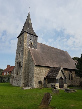A historic stone church with a prominent steeple and a clock on the tower. The building features arched windows and a sloped roof. The foreground includes a well-maintained lawn with several old gravestones, adding a rustic and tranquil atmosphere. The surrounding area has some greenery, and part of a red-brick house is visible to the side.