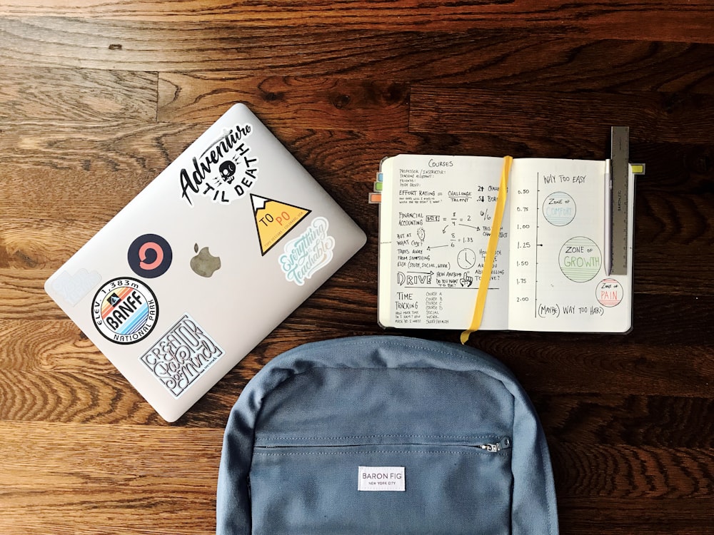 Flat-Lay-Fotografie eines blauen Rucksacks neben einem Buch und einem silbernen MacBook