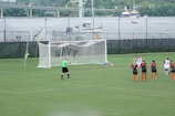 A women's soccer match taking place on a well-maintained grass field. A penalty kick is being executed, with one player in a black and white uniform scoring a goal past the goalkeeper, who is dressed in light blue and lies on the ground attempting a save. Several other players, wearing black and orange uniforms, observe the shot in action. A referee in a green shirt is positioned near the goal. In the background, there are green fences with the word 'NAVY' and a river with boats and a bridge visible.