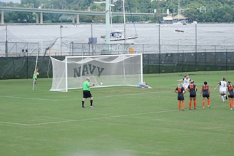 A women's soccer match taking place on a well-maintained grass field. A penalty kick is being executed, with one player in a black and white uniform scoring a goal past the goalkeeper, who is dressed in light blue and lies on the ground attempting a save. Several other players, wearing black and orange uniforms, observe the shot in action. A referee in a green shirt is positioned near the goal. In the background, there are green fences with the word 'NAVY' and a river with boats and a bridge visible.