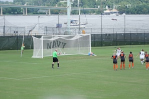 A women's soccer match taking place on a well-maintained grass field. A penalty kick is being executed, with one player in a black and white uniform scoring a goal past the goalkeeper, who is dressed in light blue and lies on the ground attempting a save. Several other players, wearing black and orange uniforms, observe the shot in action. A referee in a green shirt is positioned near the goal. In the background, there are green fences with the word 'NAVY' and a river with boats and a bridge visible.