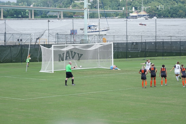 A women's soccer match taking place on a well-maintained grass field. A penalty kick is being executed, with one player in a black and white uniform scoring a goal past the goalkeeper, who is dressed in light blue and lies on the ground attempting a save. Several other players, wearing black and orange uniforms, observe the shot in action. A referee in a green shirt is positioned near the goal. In the background, there are green fences with the word 'NAVY' and a river with boats and a bridge visible.