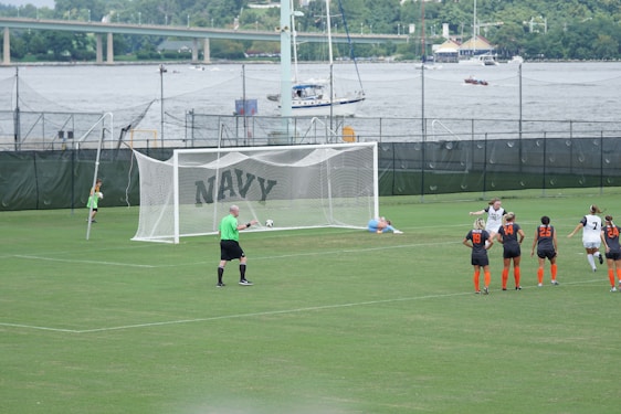 A women's soccer match taking place on a well-maintained grass field. A penalty kick is being executed, with one player in a black and white uniform scoring a goal past the goalkeeper, who is dressed in light blue and lies on the ground attempting a save. Several other players, wearing black and orange uniforms, observe the shot in action. A referee in a green shirt is positioned near the goal. In the background, there are green fences with the word 'NAVY' and a river with boats and a bridge visible.