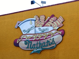 A large wall mural features an illustration of a hot dog with mustard, accompanied by a side of fries in a branded container. The background is a vibrant yellow brick wall, and the logo prominently displays the name 'Nathan's'.