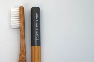 A bamboo toothbrush lies next to a cylindrical black container. The toothbrush features white bristles, and the handle exhibits a natural wood grain. The container has a smooth surface with white text and a logo.