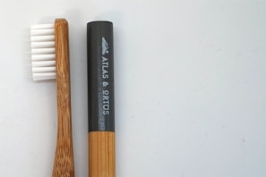 A bamboo toothbrush lies next to a cylindrical black container. The toothbrush features white bristles, and the handle exhibits a natural wood grain. The container has a smooth surface with white text and a logo.