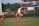 A soccer match scene with four players on the field, three wearing red uniforms and one in white. Two players are leaping to head the ball, which is in mid-air. The background shows a scoreboard displaying scores and trees in the distance.