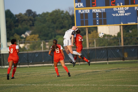 A soccer match scene with four players on the field, three wearing red uniforms and one in white. Two players are leaping to head the ball, which is in mid-air. The background shows a scoreboard displaying scores and trees in the distance.