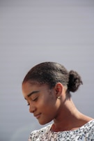A woman with a serene expression is looking downwards. She has her hair neatly pulled back into a bun and is wearing a patterned black and white top. The background is plain and softly lit, giving prominence to her calm demeanor.