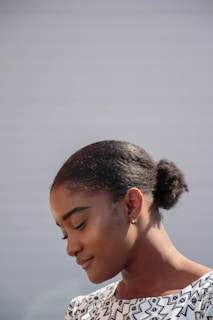 A woman with a serene expression is looking downwards. She has her hair neatly pulled back into a bun and is wearing a patterned black and white top. The background is plain and softly lit, giving prominence to her calm demeanor.