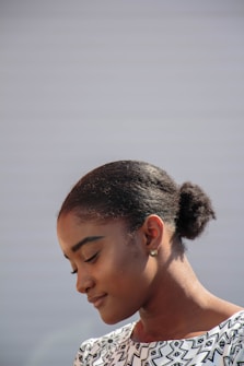 A woman with a serene expression is looking downwards. She has her hair neatly pulled back into a bun and is wearing a patterned black and white top. The background is plain and softly lit, giving prominence to her calm demeanor.