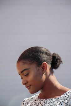 A woman with a serene expression is looking downwards. She has her hair neatly pulled back into a bun and is wearing a patterned black and white top. The background is plain and softly lit, giving prominence to her calm demeanor.