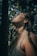 A person stands outdoors with the head tilted upward, partially illuminated by dappled sunlight filtering through the trees. Dirt is visible on the face and shoulders, suggesting an interaction with nature or wilderness. The expression is contemplative or pensive as they gaze upward.