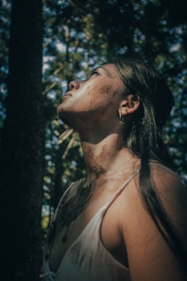 A person stands outdoors with the head tilted upward, partially illuminated by dappled sunlight filtering through the trees. Dirt is visible on the face and shoulders, suggesting an interaction with nature or wilderness. The expression is contemplative or pensive as they gaze upward.