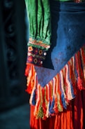 A colorful garment with intricate embroidery and patterns is visible. The fabric features vibrant colors including green, blue, red, and orange, with detailed decorative fringes and buttons.