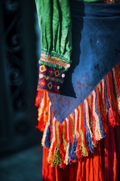 A colorful garment with intricate embroidery and patterns is visible. The fabric features vibrant colors including green, blue, red, and orange, with detailed decorative fringes and buttons.