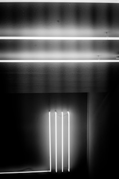 Two horizontal and three vertical glowing white neon tubes create a minimalist geometric pattern against a dark background.