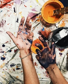 Hands covered in multicolored paint with paint buckets nearby, resting on a paint-splattered tarp.