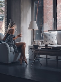 A person sits on a couch, wrapped in a cozy blanket, holding a cup and looking out a large window. The room has a calm and minimalist aesthetic, featuring a table with a lamp and some decorative pillows. Soft natural light streams in through the window, creating a warm and tranquil atmosphere.