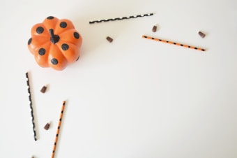 An orange pumpkin with black polka dots is placed on a white background. Around the pumpkin, there are straws with black and orange stripes and dots, along with small brown chocolate pieces scattered in a minimalistic arrangement.