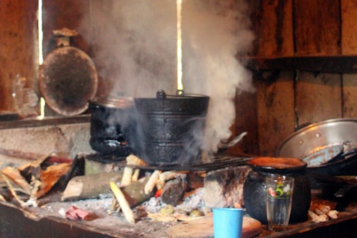 An indoor rustic kitchen scene with wooden walls and a stone stove is visible. Smoke rises from a black cooking pot set on top of a wood fire. Nearby, various cooking utensils and containers, including a blue plastic cup and a metal pot lid, are scattered.
