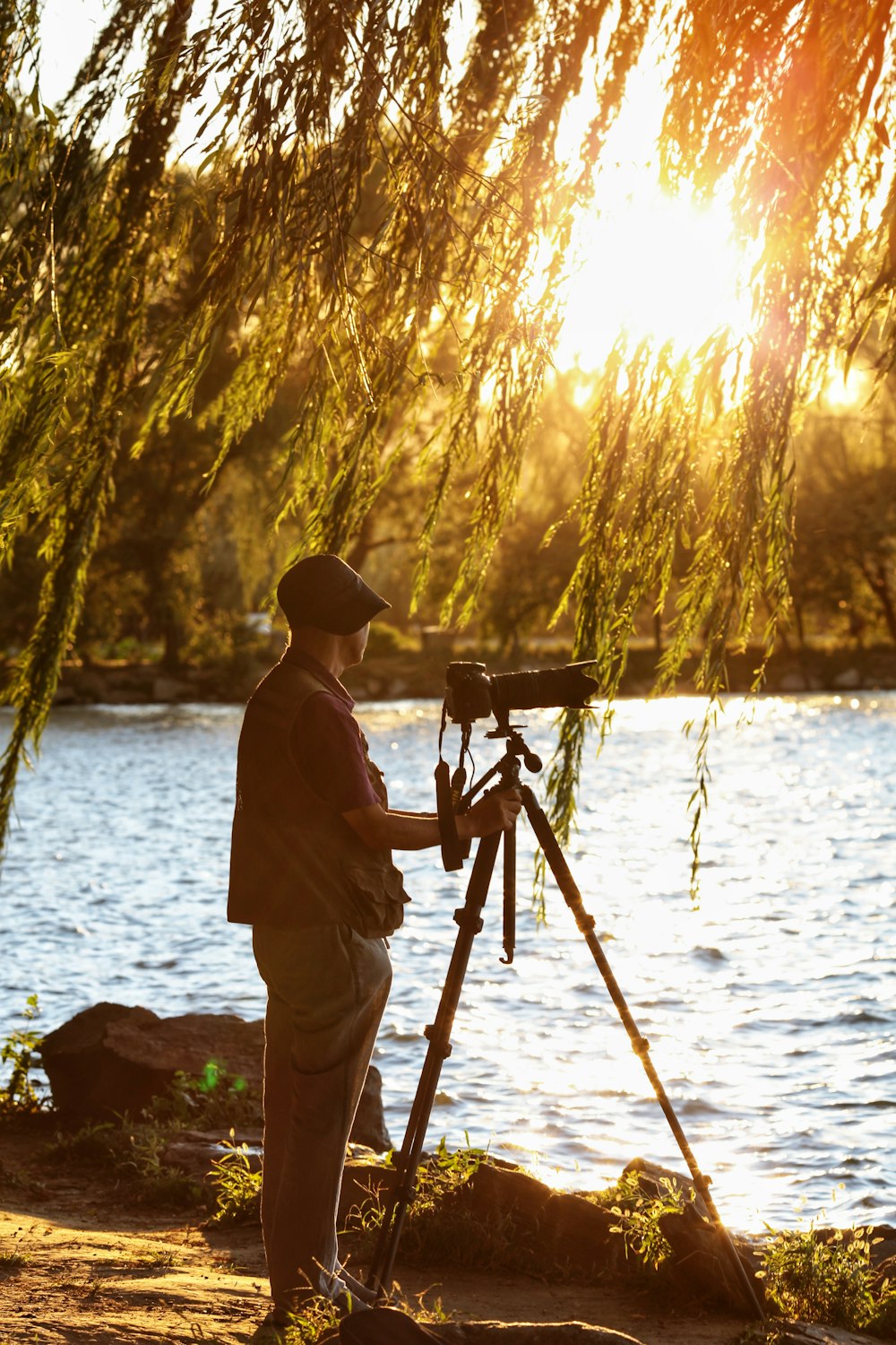 man standing beside DSLR camera with stand near body of water during sunset