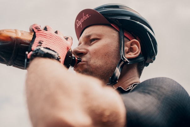 A man wearing a bicycle helmet and drinking from a bottle. He has a determined and focused expression. He is also wearing red and black cycling gloves.