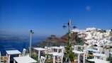 An outdoor seating area with white tables and green chairs is situated on a terrace overlooking a stunning view. In the background, a cliffside is adorned with iconic white buildings common to Greek islands. The sea is visible in the distance under a clear blue sky, contributing to the serene ambiance.