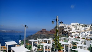 An outdoor seating area with white tables and green chairs is situated on a terrace overlooking a stunning view. In the background, a cliffside is adorned with iconic white buildings common to Greek islands. The sea is visible in the distance under a clear blue sky, contributing to the serene ambiance.