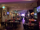 A warmly lit restaurant interior features several tables and chairs, with patrons dining. Framed art and television screens displaying images decorate the walls. The space is softly illuminated by decorative hanging lamps, and a stage with musical equipment is visible at the far end.