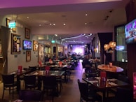 A warmly lit restaurant interior features several tables and chairs, with patrons dining. Framed art and television screens displaying images decorate the walls. The space is softly illuminated by decorative hanging lamps, and a stage with musical equipment is visible at the far end.
