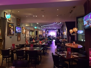 A warmly lit restaurant interior features several tables and chairs, with patrons dining. Framed art and television screens displaying images decorate the walls. The space is softly illuminated by decorative hanging lamps, and a stage with musical equipment is visible at the far end.