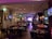 A warmly lit restaurant interior features several tables and chairs, with patrons dining. Framed art and television screens displaying images decorate the walls. The space is softly illuminated by decorative hanging lamps, and a stage with musical equipment is visible at the far end.