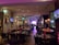 A warmly lit restaurant interior features several tables and chairs, with patrons dining. Framed art and television screens displaying images decorate the walls. The space is softly illuminated by decorative hanging lamps, and a stage with musical equipment is visible at the far end.