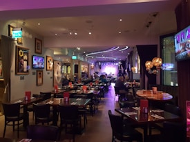 A warmly lit restaurant interior features several tables and chairs, with patrons dining. Framed art and television screens displaying images decorate the walls. The space is softly illuminated by decorative hanging lamps, and a stage with musical equipment is visible at the far end.