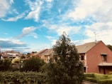 A suburban neighborhood with red-roofed houses under a vibrant sky filled with scattered clouds. Lush greenery, including bushes and trees, surrounds the buildings, providing a tranquil atmosphere.