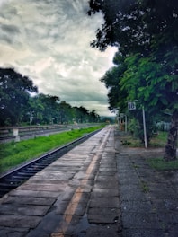 A railway platform stretches into the distance, bordered by lush green trees and a cloudy sky overhead. The platform is wet, suggesting recent rain. There are signposts along the side, and tracks run parallel to a strip of grass.