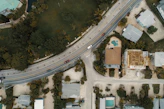 An aerial view of a suburban area featuring a curving road with several cars, surrounded by residential houses and lush greenery. One house has an unfinished roof, indicating construction work, while another has a visible swimming pool in the backyard. The arrangement of houses and roads is complemented by the presence of palm trees and other vegetation.