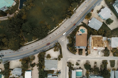 An aerial view of a suburban area featuring a curving road with several cars, surrounded by residential houses and lush greenery. One house has an unfinished roof, indicating construction work, while another has a visible swimming pool in the backyard. The arrangement of houses and roads is complemented by the presence of palm trees and other vegetation.